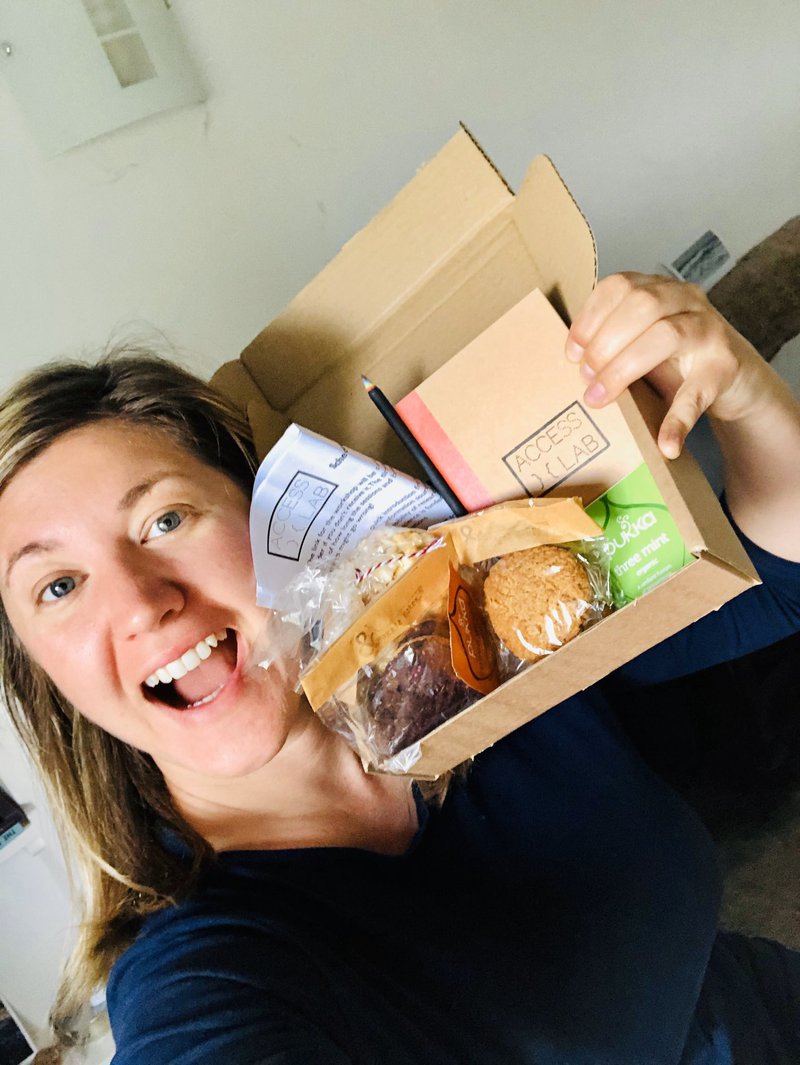 Smiling person holding a small box containing biscuits, teabags, notebook, pencil and a piece of printed paper